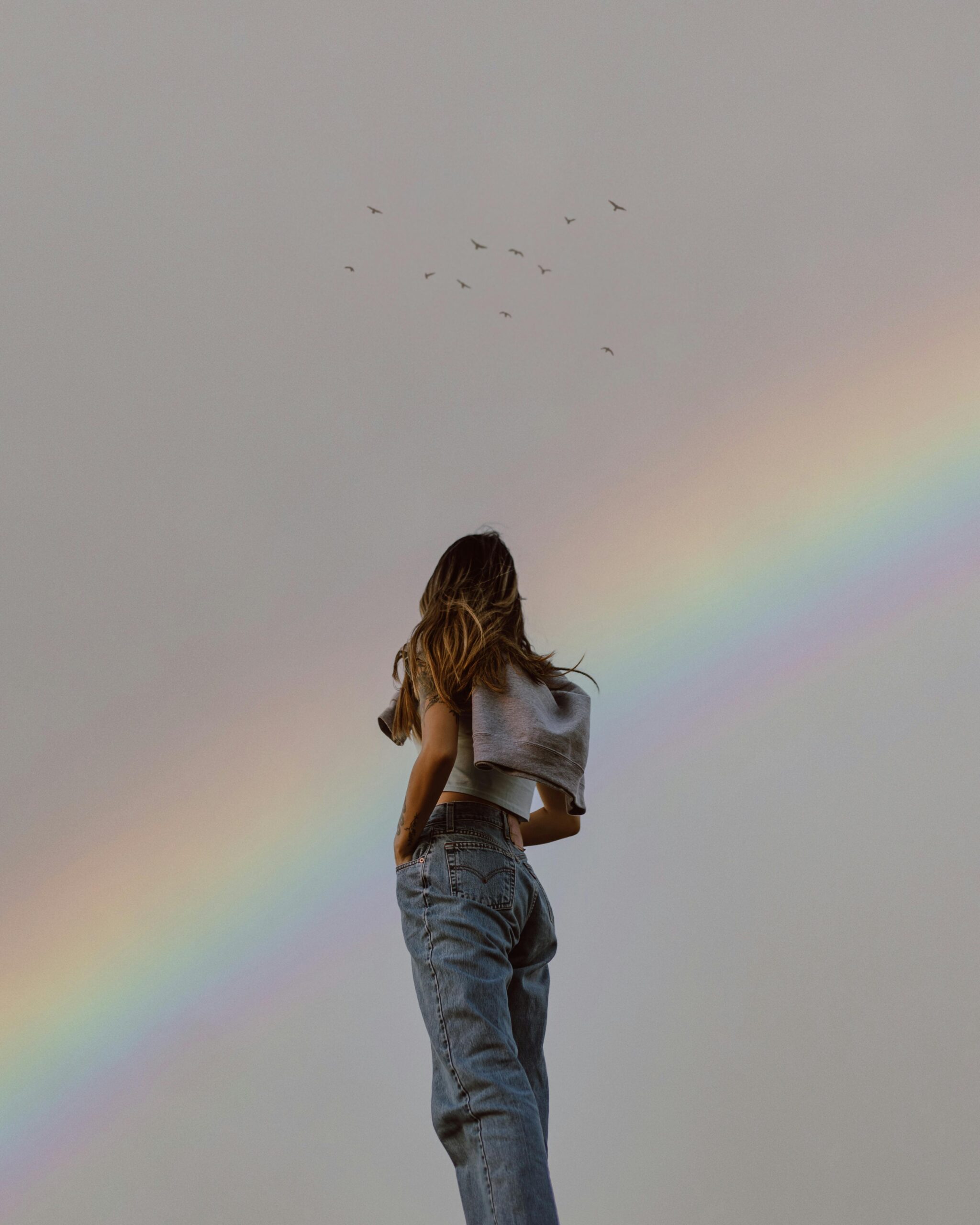 young woman standing with her hands in her jeans pockets and looking out at a muted rainbow